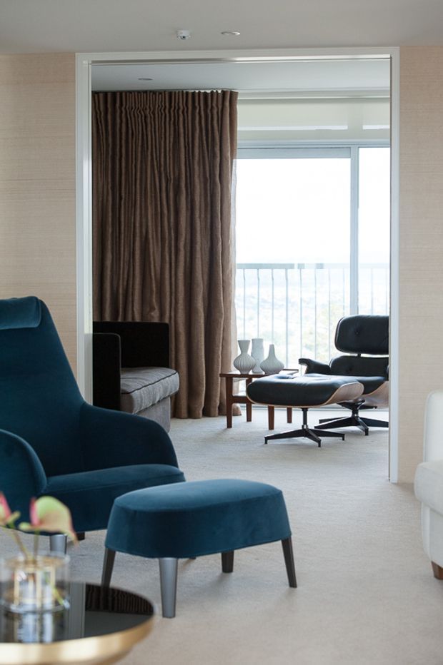 A refined contemporary living room viewed through a wide opening, with deep blue upholstered lounge chairs and ottoman in the foreground, a black-and-tan mid-century-style lounge chair and footstool beyond, a small timber coffee table with sculptural vases, full-height brown curtains, pale textured walls, and soft light-coloured carpet. Large glazing opens to a balcony, creating a bright, calm interior.