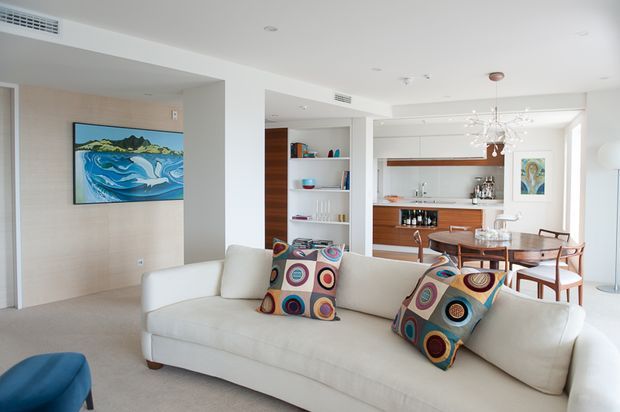 Bright open-plan living and dining area with a white upholstered sofa in the foreground, colourful geometric cushions, pale neutral walls and carpet, and a timber-accented built-in bar or kitchen joinery behind the dining table. The room includes recessed ceiling lighting, a contemporary branching pendant over the dining area, built-in shelving, and coastal artwork.