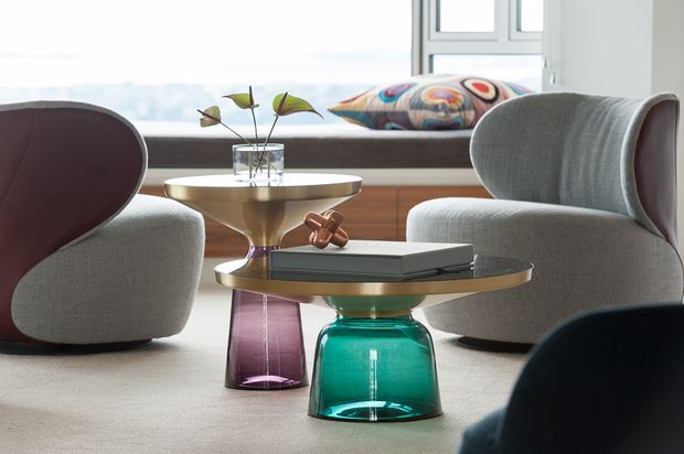 A refined contemporary living room vignette with sculptural upholstered lounge chairs, two low round coffee tables featuring brass-toned tops and colourful glass pedestal bases, and a built-in window seat with a patterned cushion. The space is softly lit and uses a muted neutral palette with jewel-toned accents.