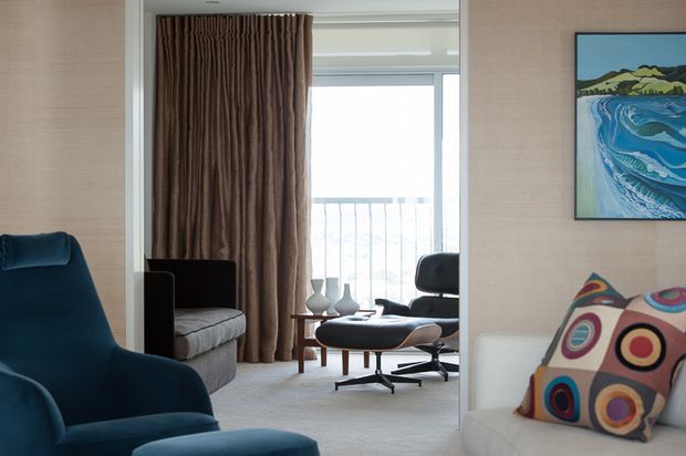 A refined living room with a blue upholstered lounge chair in the foreground, a white sofa with a colourful geometric cushion, and a mid-century style lounge chair and ottoman near full-height glazing. Brown drapery, pale textured wall finishes, carpeted flooring, and a framed coastal artwork create a warm, curated interior.