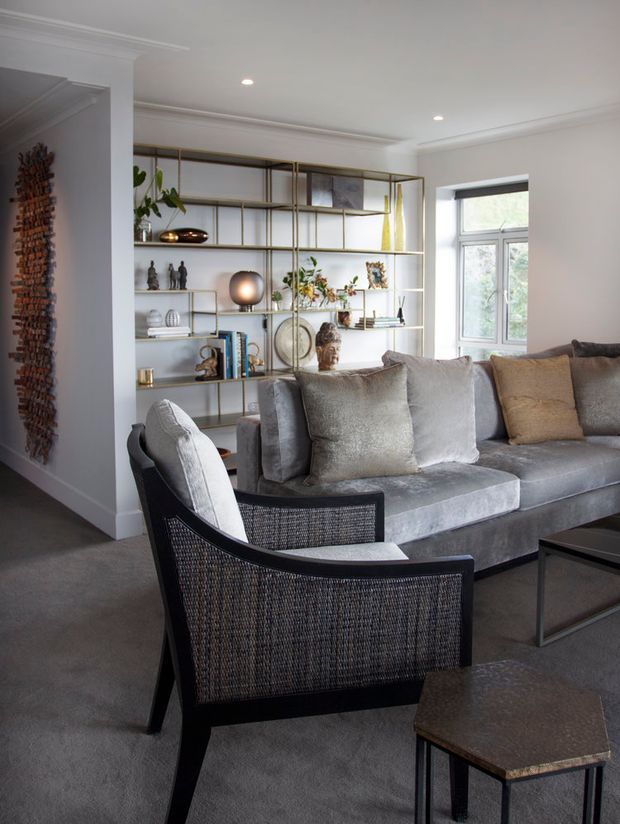 A refined living room with a large grey upholstered sectional sofa, a black woven lounge chair, and a dark geometric coffee table. A full-height brass-toned open display shelving unit spans the back wall, styled with books, artwork, sculptures, ceramics, and plants. The room has white walls with decorative cornices, recessed ceiling lights, grey carpet, and a large window bringing in natural light. A textured wall hanging is visible in the adjacent area.