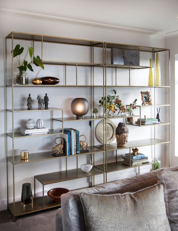 A refined living room featuring a large floor-to-ceiling open metal shelving installation with warm brass-toned framing and dark shelves. The shelves display books, sculptures, ceramics, plants, artwork and decorative objects, with a sofa in the foreground. White walls with traditional crown moulding, soft neutral upholstery, and warm decorative lighting create an elegant, curated atmosphere.