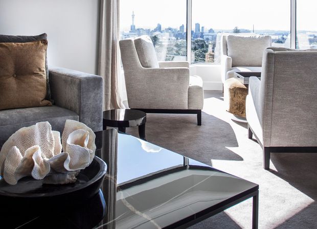 Bright, refined living room with floor-to-ceiling windows overlooking a city skyline. The room features a large neutral sectional sofa, upholstered lounge chairs, a glass-topped black coffee table, dark side tables, floor-length curtains, and soft beige carpeting. The composition is contemporary and polished, with strong natural daylight and an expansive urban view.