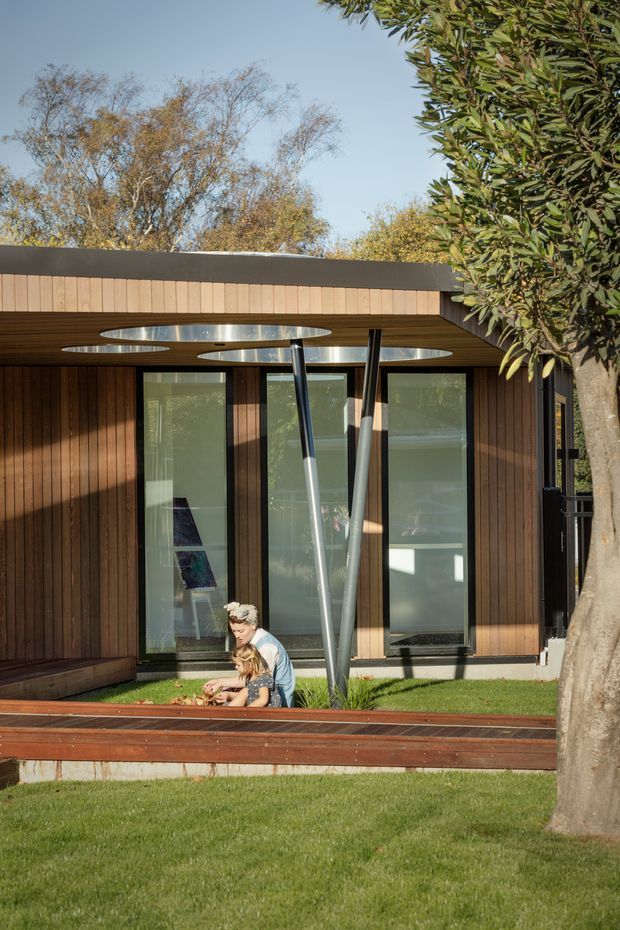 Exterior view of a low-slung contemporary building with warm vertical timber cladding, expansive glazed doors and windows, and a projecting flat roof canopy featuring circular skylight openings. Sculptural angled steel supports frame the entry or covered outdoor area. The building sits beside a timber deck and manicured lawn, with mature trees surrounding it.