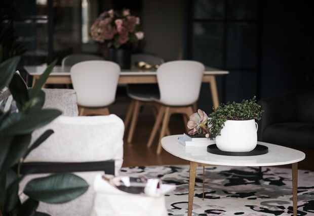 A moody contemporary living and dining interior with a pale upholstered sofa in the foreground, a round white coffee table with potted plants, and a patterned area rug. A light timber dining table and sculptural upholstered dining chairs sit behind the living area, framed by dark walls and glazing, with indoor plants and floral arrangements adding natural texture.
