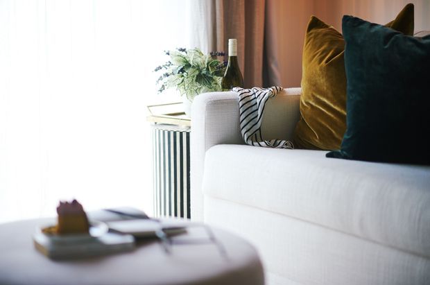Close-up interior view of a softly styled living room, featuring a light upholstered sofa with mustard and deep teal cushions, a striped throw, a round coffee table, and a decorative fluted side table with greenery and a wine bottle. Warm curtains and bright window light create a refined, inviting atmosphere.