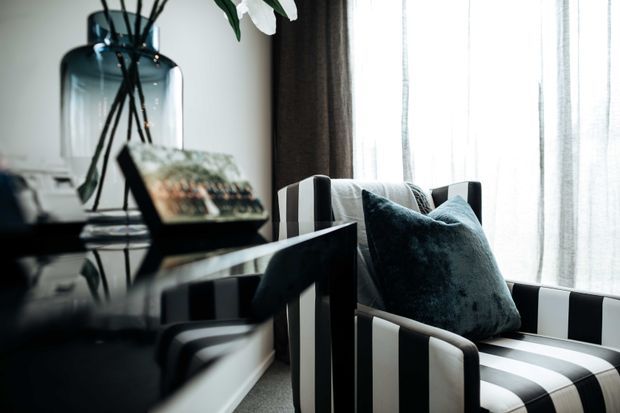 A refined sitting corner featuring a black-and-white vertically striped armchair beside a glossy dark console or side table. A deep teal velvet cushion adds contrast, while sheer curtains diffuse daylight from a large window. Decorative glass and greenery sit on the surface, creating a polished, high-contrast interior vignette.