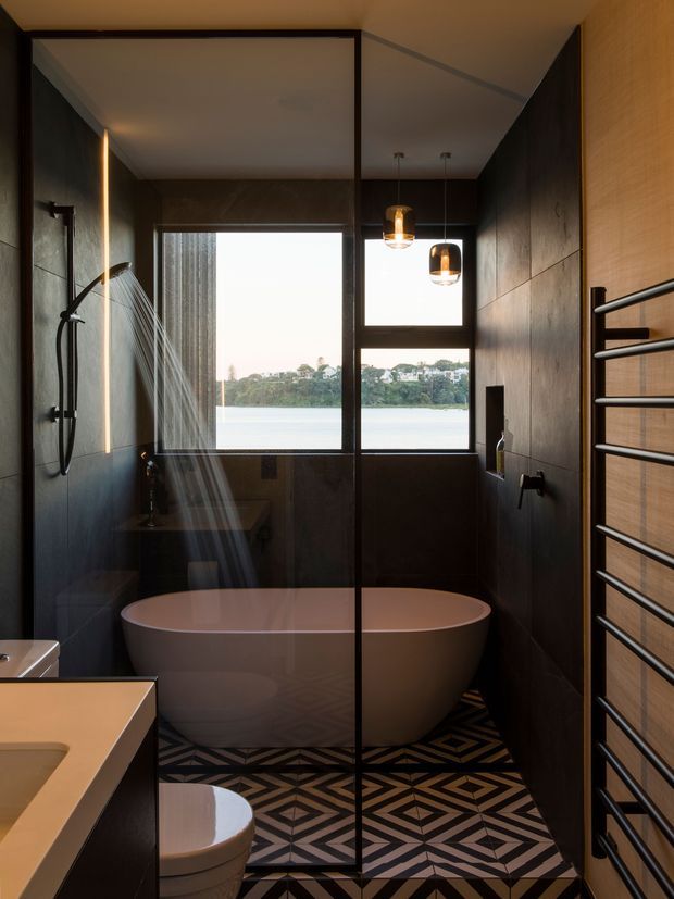 A compact, moody contemporary bathroom with a freestanding white bathtub positioned beside a frameless glass shower enclosure. Dark wall panels, geometric patterned floor tiles, a wall-mounted towel warmer, pendant lights, and large black-framed windows overlooking a body of water create a dramatic, refined interior.