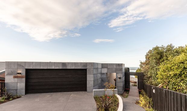 Exterior view of a contemporary single-storey residence focused on a dark double garage door set within a rectilinear grey masonry or concrete-block facade. A concrete driveway and narrow paved walkway lead past a curved raised garden bed, with dark fencing and established planting at the side. The flat-roofed form and strong geometric composition are prominent.
