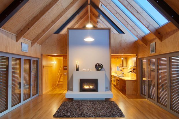 Warm timber-lined open-plan interior with a dramatic vaulted ceiling and exposed timber rafters. A central freestanding fireplace with a pale rendered surround divides the living area from a visible kitchen behind it. Skylights run along the pitched roof, while timber-framed louvered doors and windows line both sides. The room has polished timber flooring, a dark textured rug, recessed cabinetry, and pendant lighting.