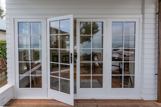 Exterior view of a white weatherboard-clad facade with a set of multi-pane glazed French doors opening onto a timber deck. Through the doors, a small furnished interior with chairs and a table is visible, along with reflections of the surrounding landscape. The image has a bright coastal, traditional character.