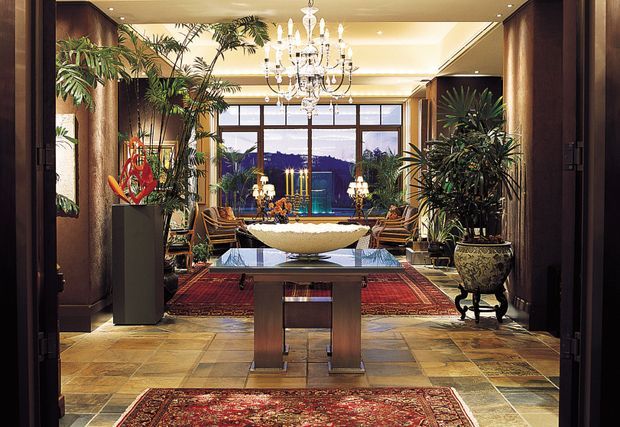 A richly appointed formal living room or reception space viewed through wide dark timber doors. The room features large indoor palms and other potted plants, a dramatic crystal chandelier, a sculptural central console or table, ornate seating, Persian-style rugs, stone or tiled flooring, dark timber wall panelling, and large windows overlooking a distant landscape. The overall atmosphere is luxurious, warm, and eclectic with strong traditional influences.