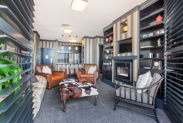A stylish, compact living room with a dark built-in shelving and fireplace wall, extensive vertical and horizontal window blinds, striped wall detailing, leather armchairs, a patterned lounge chair, and a cowhide-topped coffee table. The room has a layered, refined interior with strong contrast between dark joinery, warm leather, and light upholstery.