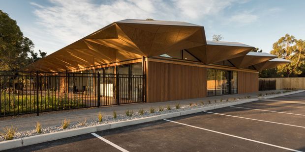 Exterior view of a contemporary single-storey building with faceted, angular roof planes and extensive glazing. Warm timber-look vertical wall cladding and dark metal framing define the facade, with a fenced landscaped edge and marked parking area in front.