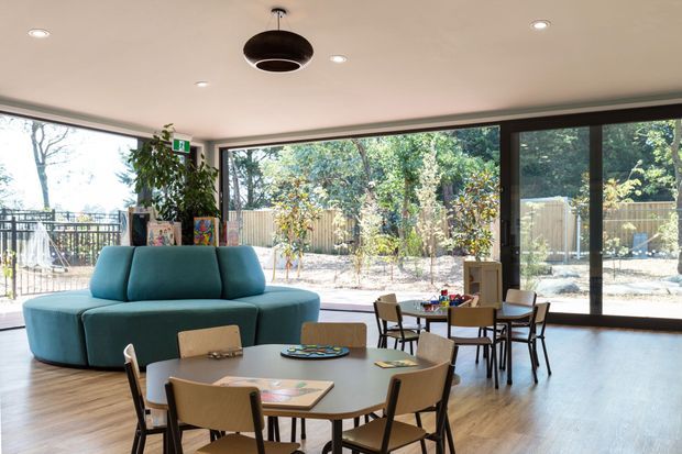 Bright, open-plan communal living and dining space with floor-to-ceiling black-framed glazing overlooking a landscaped outdoor area. A curved teal sofa, two round dining tables with timber chairs, indoor plants, artwork, and a suspended dark pendant create a playful yet contemporary interior.