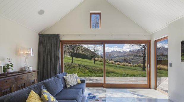 Bright, high-gabled living room with a large timber-framed picture window and glazed door opening onto a deck and landscaped rural mountain setting. The room features white painted walls and ceiling boards, a blue sectional sofa, timber sideboard, neutral curtains, and a patterned area rug. The expansive glazing and vaulted ceiling create an airy connection to the outdoors.