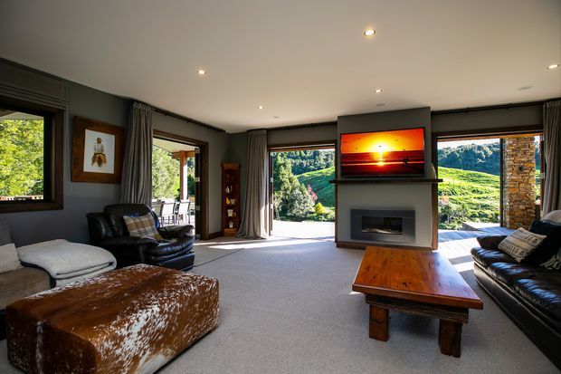 A spacious contemporary living room with dark painted walls, recessed ceiling lighting, expansive windows and glazed doors opening to a scenic green landscape. The room features a central fireplace with a wall-mounted television above it, black leather seating, a large cowhide-covered ottoman, a substantial rustic timber coffee table, and floor-length curtains.