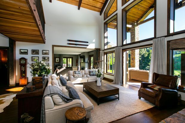 Spacious double-height living room with expansive black-framed glazing, warm timber ceiling and exposed beams, stone accents, and multiple neutral upholstered sofas arranged around a substantial timber coffee table. A leather armchair, traditional grandfather clock, framed artwork, soft curtains, and layered furnishings create a warm, refined interior with strong indoor-outdoor connection.