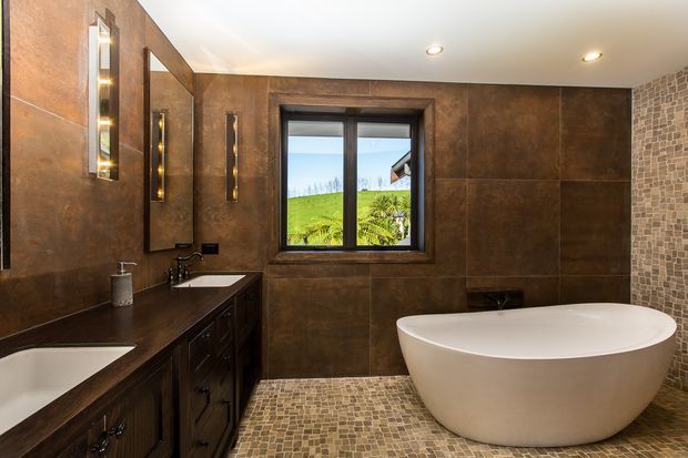 A refined bathroom with a freestanding oval bathtub, double vanity, dark timber cabinetry, large brown stone-look wall panels, mosaic floor tiles, a textured mosaic feature wall, black-framed window, and warm recessed and wall lighting. A green landscape is visible outside the window.