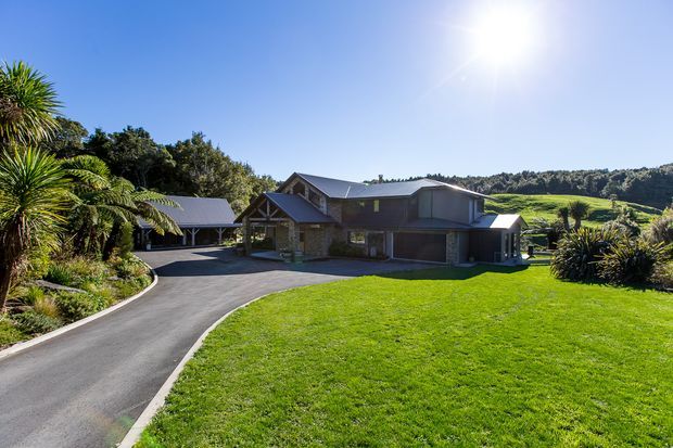 A broad exterior view of a contemporary multi-level house set within a landscaped, semi-rural site. The dark-clad home has a complex pitched roofline, extensive glazing, stonework at the lower facade, and a covered carport or outbuilding to the left. A curved asphalt driveway crosses the manicured lawn, with mature native-style planting and rolling green hills beyond.