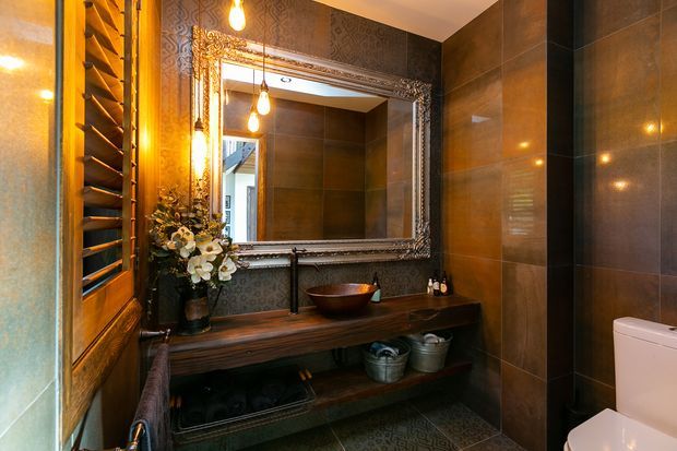 A richly detailed, dark-toned bathroom with large brown wall tiles, patterned feature wallpaper, a timber vanity with open shelving, black fixtures, and a decorative framed mirror. Warm pendant and wall lighting create an intimate, luxurious atmosphere. A toilet and shower enclosure are visible on the right, while a louvred window or screen appears on the left.