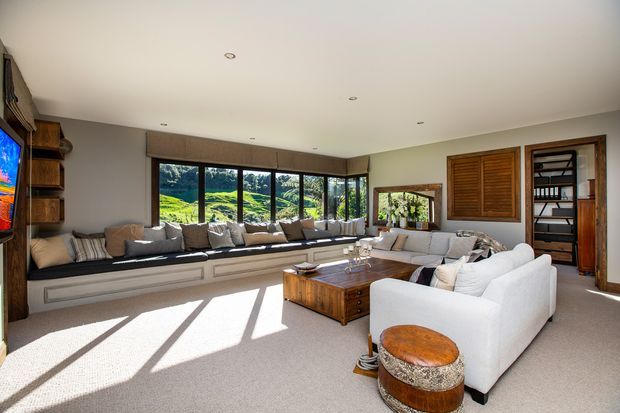 Bright, spacious living room with a long built-in window seat, expansive black-framed glazing overlooking a green landscape, white upholstered sofas, a timber coffee table, and warm timber shutter and shelving details. The room has recessed ceiling lights, soft carpet flooring, and a relaxed contemporary feel.