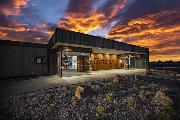 Exterior view of a contemporary commercial building at sunset, with a dark charcoal metal-clad facade, projecting entrance canopy, recessed downlights, extensive glazing, and a warm timber-lined entry wall bearing commercial signage. The building is set within a gravelled, sparsely planted landscape with sculptural rocks.