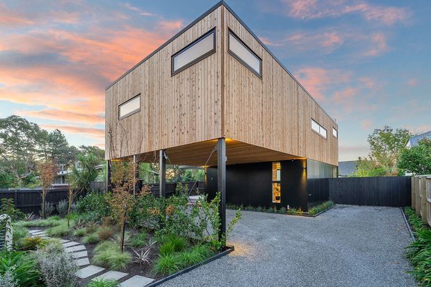 Exterior view of a contemporary elevated timber-clad house at dusk. The upper floor has a steep gable-like form with narrow horizontal windows, while the dark lower level is set above an open carport or undercroft supported by slender posts. A gravel driveway, landscaped garden, stepping-stone path, and dark fencing surround the home.