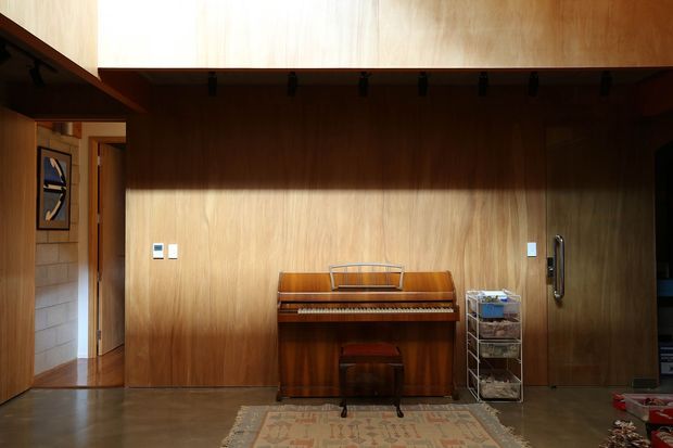 Interior view of a compact timber-lined room with a freestanding upright piano against a full-height wood wall. A small patterned rug, piano stool, metal storage trolley, framed artwork, and doorway are visible. The space has a warm, restrained mid-century character with a dark polished floor and simple functional detailing.