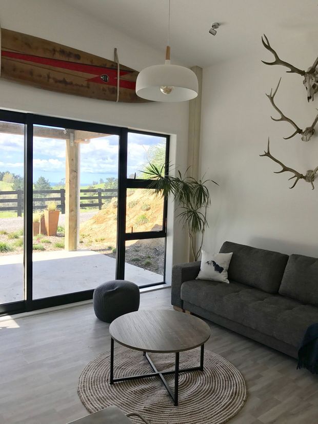 Bright contemporary living room with a dark grey sofa, round wood-look coffee table, textured circular rug, pendant light, indoor plant and decorative mounted antlers. Large black-framed glazed doors open to an outdoor patio and landscaped setting, while a vintage-style wooden board adds a rustic accent above the glazing.