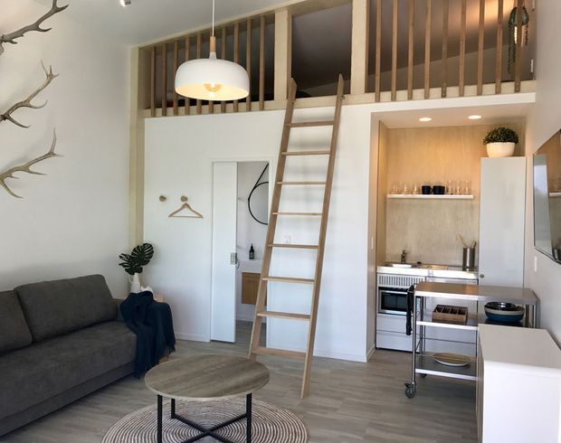 Compact open-plan studio interior with a living area and kitchenette beneath a loft sleeping platform. The room features a timber ladder, vertical timber balustrade, pale wood cabinetry and wall paneling, light wood-look flooring, a dark sofa, round coffee table, and contemporary pendant lighting.