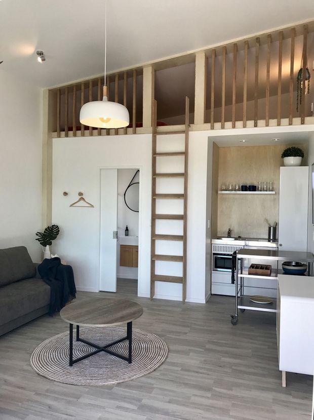 Bright compact open-plan apartment interior with a living area, kitchenette, and loft sleeping platform. A timber ladder leads to the elevated mezzanine, which is enclosed by vertical timber balusters. The space features pale walls, grey timber-look flooring, white cabinetry, a grey sofa, round coffee table, pendant light, and a woven circular rug.