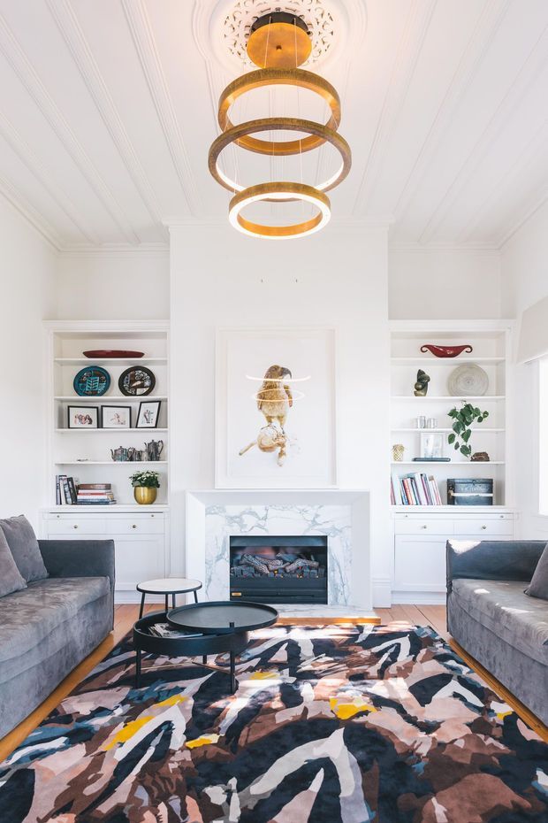 Bright, refined living room with white built-in shelving flanking a marble-faced fireplace, decorative wall art, grey sofas, black round coffee tables, and a bold abstract patterned rug. The room features ornate moulded ceiling detailing and a dramatic tiered brass-and-bronze ring pendant light.