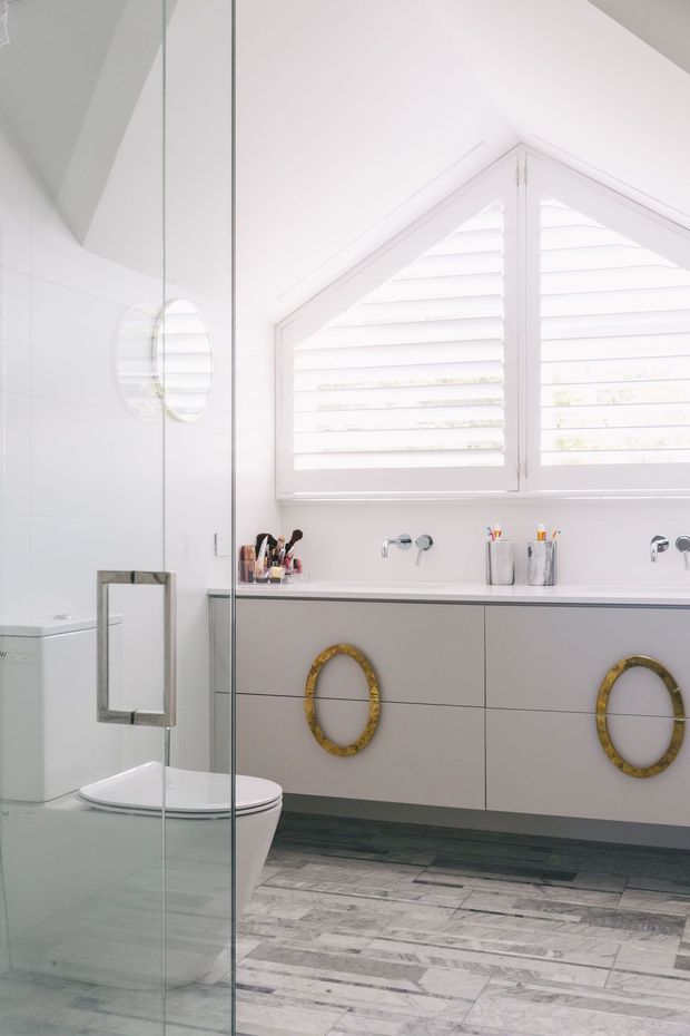 Bright contemporary bathroom with a frameless glass shower enclosure, wall-mounted double vanity, white fixtures, and a large angled window fitted with white plantation shutters. The vanity has pale grey flat-front drawers with distinctive round brass-gold pulls, a slim dark benchtop, and the floor is finished in elongated grey-and-white stone-look tiles.