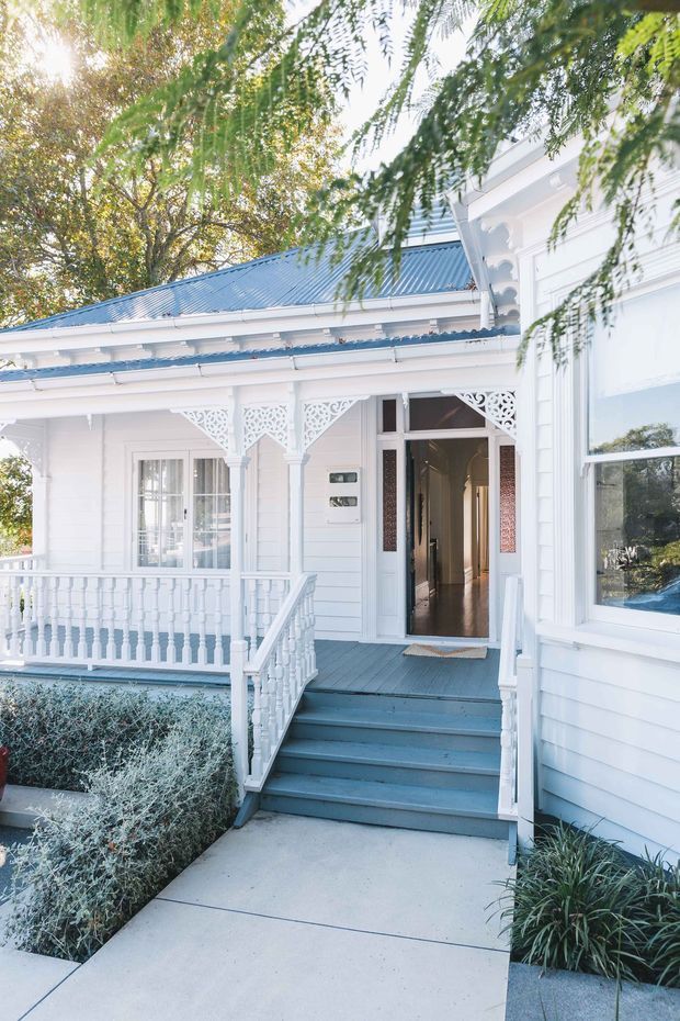 Bright exterior view of a white timber-clad heritage-style house entrance, featuring a blue-grey corrugated metal roof, ornate decorative fretwork, turned porch balusters, a covered veranda, front steps, and a concrete garden path. Mature planting partially frames the facade.