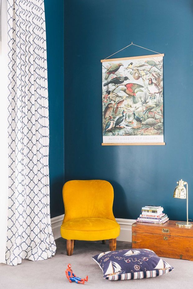 A richly coloured interior corner featuring deep teal walls, white patterned curtains, and a mustard-yellow upholstered armchair. A vintage bird illustration hangs above a wooden chest used as a side table, with a brass desk lamp and stacked books. A patterned floor cushion and toy sit on the carpet, creating an eclectic, playful reading or sitting nook.