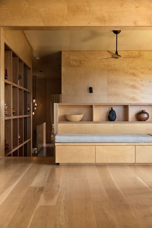 Warm, timber-lined interior featuring a built-in window seat or bench with pale upholstered cushion, integrated drawers, and open display shelving containing ceramic vessels. A full-height timber screen and shelving unit separates the seating area from a darker hallway, while a ceiling fan and pendant lights add functional lighting and ventilation. The space has a cohesive natural-wood fit-out and timber flooring.