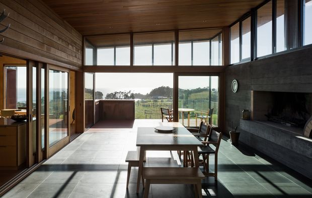 A spacious, light-filled open-plan living and dining area with extensive floor-to-ceiling glazing overlooking a rural landscape. The room features a long timber dining table with wooden chairs, a large dark masonry fireplace, warm timber-lined walls and ceiling, dark-framed glazing, and large pale stone or concrete floor tiles. A glazed opening on the left connects visually to another interior space.