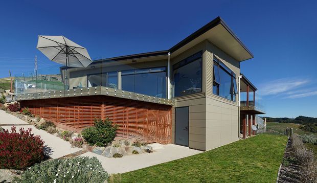Contemporary single-storey house on a sloped rural site, featuring dark horizontal cladding, extensive glazing, a broad roof overhang, timber screening, and an elevated terrace with an outdoor umbrella. Landscaped planting and lawn surround the building, with open countryside visible beyond.