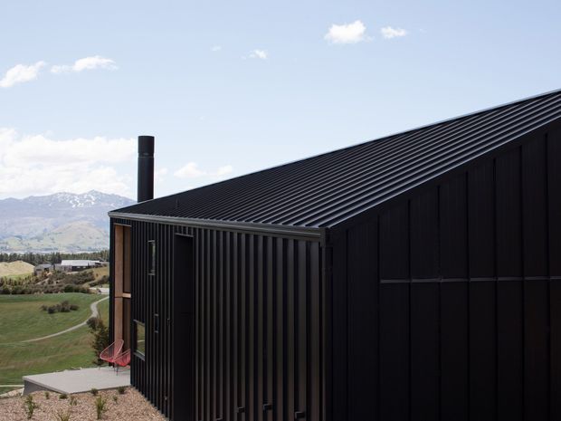 Exterior view of a dark, minimalist single-storey building with a steeply pitched ribbed metal roof and vertically clad dark facade. A chimney rises near the roof edge, while tall narrow glazing and a small terrace are visible on the side. The building sits in an open rural or mountainous landscape with distant snow-capped hills.
