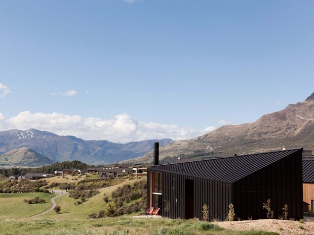 A modern dark-clad building with a sharply pitched metal roof sits in a landscaped rural valley, with large mountains and blue sky in the background. The building has vertical ribbed cladding, a prominent chimney, and expansive glazing or openings along one side.