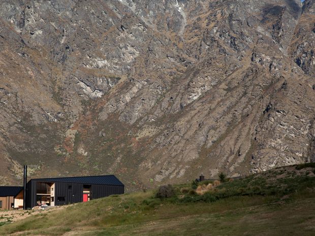 A low, modern dark-clad building sits in a rugged alpine mountain landscape. The structure has a shallow pitched roof, large glazed openings, and timber-toned elements, with steep exposed rocky mountains dominating the background and grassy terrain in the foreground.