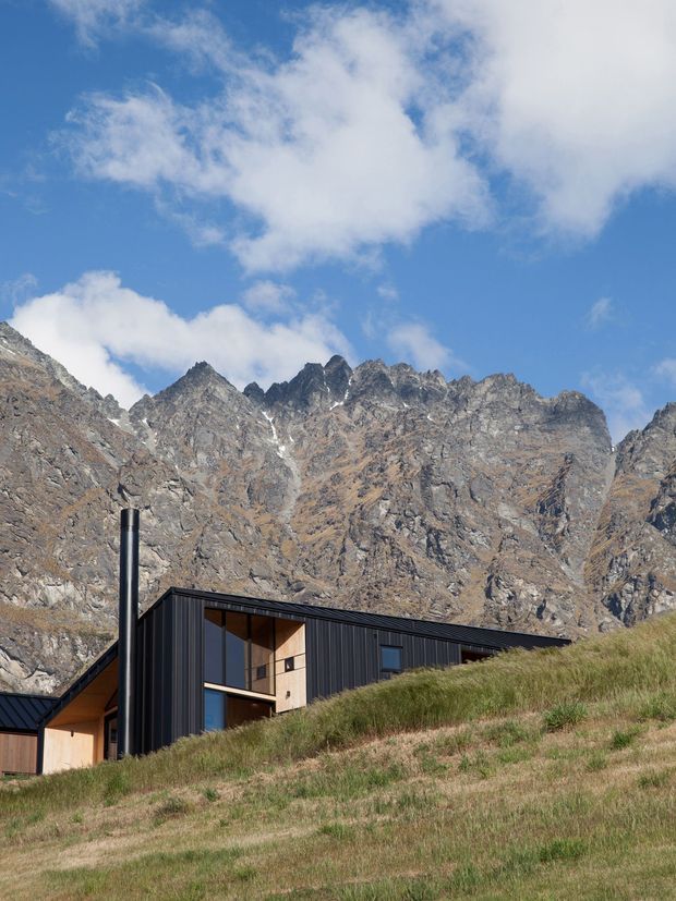 A contemporary dark-clad mountain dwelling sits partially embedded in a grassy alpine slope. The building has crisp angular rooflines, extensive timber-framed glazing, warm timber interior or soffit surfaces, and a prominent black flue. Rugged, jagged snow-dusted mountains rise immediately behind it beneath a bright blue sky with scattered clouds.