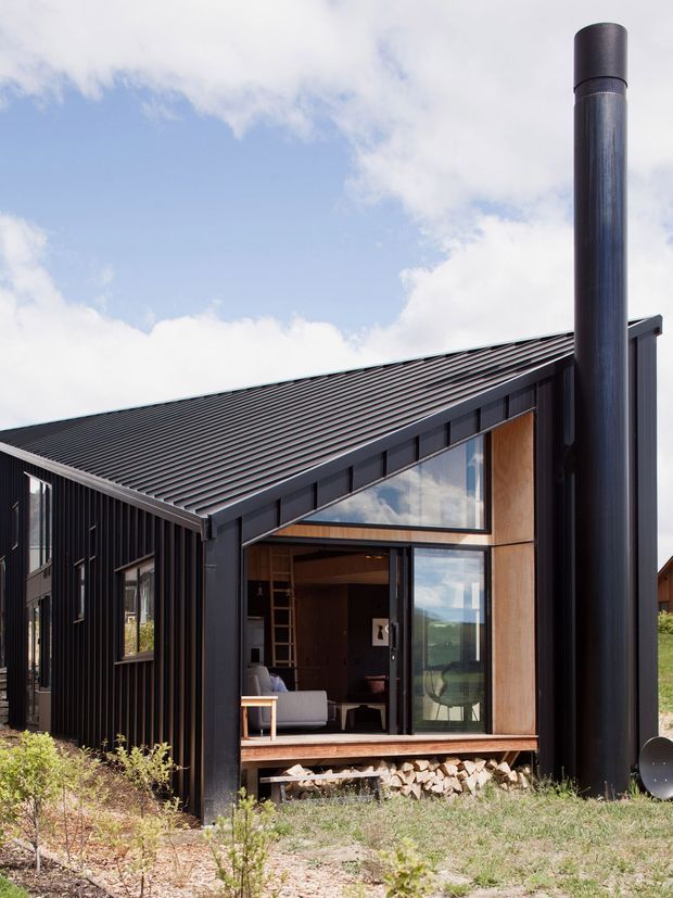Exterior view of a compact contemporary cabin with a steeply sloping corrugated metal roof, dark vertical cladding, large glazed gable frontage, warm timber wall panels, and a prominent cylindrical black flue. A living area is visible through the glazing, with firewood stored beneath the raised structure. The building sits in a sparse natural landscape beneath a partly cloudy sky.
