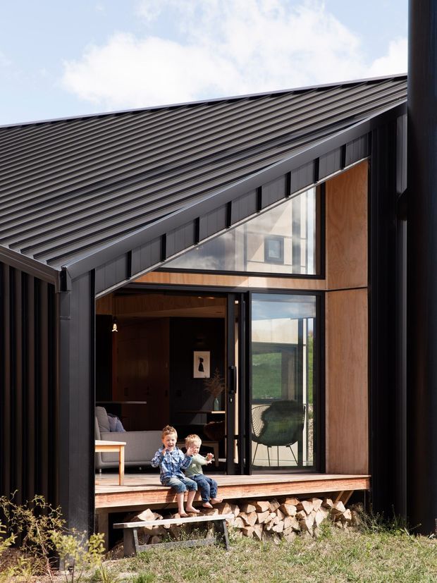 Exterior view of a contemporary dark-clad house with a steep gabled roof, extensive glazing, and a timber-lined recessed entry or deck. The open glass doors reveal a warm living area, while stacked firewood sits beneath the raised timber platform. The image also shows a triangular clerestory window and strong black structural framing.