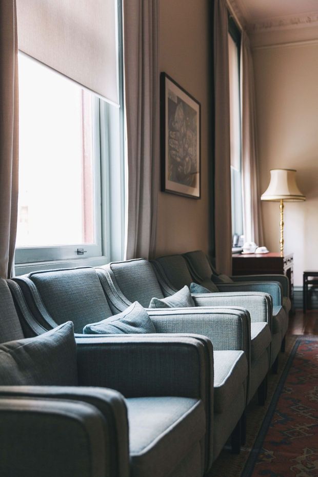 A refined sitting room with a row of matching blue-grey upholstered armchairs arranged along tall windows. The room features full-height curtains, a patterned rug, framed artwork, polished timber flooring, a traditional floor lamp, and ornate ceiling cornice detailing. Natural light creates a calm, elegant atmosphere.