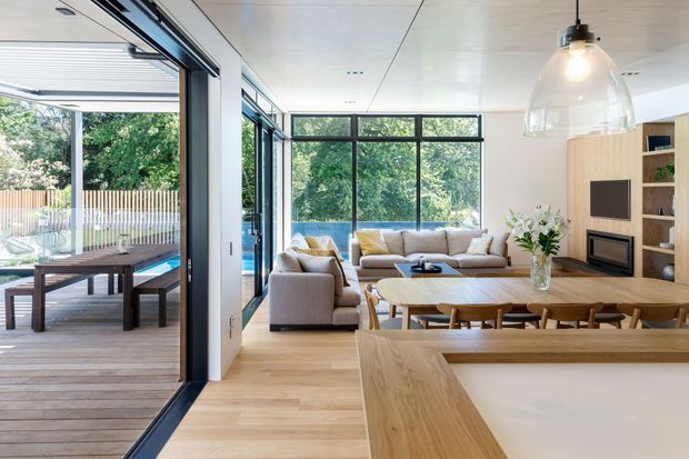 Bright open-plan living area with a contemporary timber kitchen island in the foreground, a dining table and chairs, and a comfortable lounge beyond. Floor-to-ceiling black-framed glazing opens onto a timber deck and greenery, while built-in timber cabinetry surrounds a wall-mounted television and fireplace. A large glass pendant light hangs above the kitchen area.