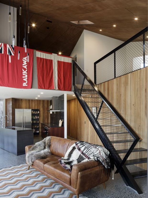 Open-plan loft-style interior combining a kitchen, living area and dramatic metal staircase. The space features a dark timber ceiling with recessed lighting, vertical timber wall panelling, black wire-mesh stair balustrades, polished concrete or terrazzo-like flooring, a dark kitchen island, and a brown leather sofa. A large red-and-white fabric banner hangs along the mezzanine edge.