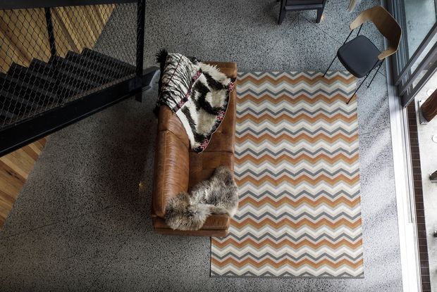Top-down view of a compact industrial-style living area with a cognac brown leather sofa, layered fur and patterned throws, a multicoloured zigzag area rug, and a simple wooden-framed chair. The space has a polished speckled concrete floor, black metal mesh architectural screening, and strong natural light from large windows or glazed doors.