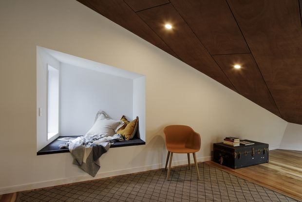A warm, minimalist attic-like interior with a sloping dark timber-paneled ceiling, recessed spotlights, white walls, and a low built-in window seat filled with cushions and a throw. A simple caramel lounge chair sits beside a dark vintage trunk used as a low table, while patterned flooring and exposed timber floorboards add texture.